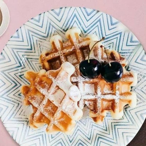 Two golden waffles dusted with powdered sugar and topped with two dark cherries on a patterned plate.